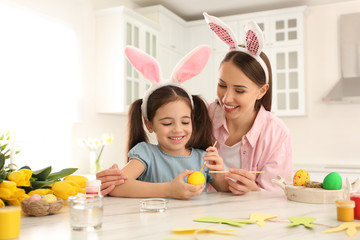 Happy mother and daughter with bunny ears headbands painting Easter egg in kitchen