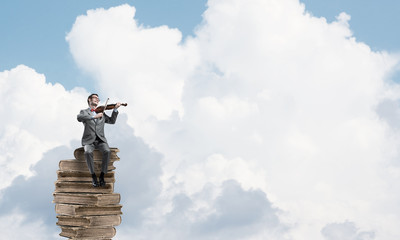 Handsome violinist in blue day sky on pile of books play his melody