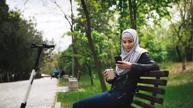 Young moderm attractive Muslim woman wearing hijab sitting in park and listening to music with smartphone and cup of coffee in her hands