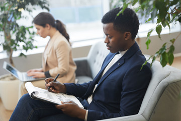 High angle portrait of successful African businessman reading document while relaxing in comfortable chair at office building hall, copy space