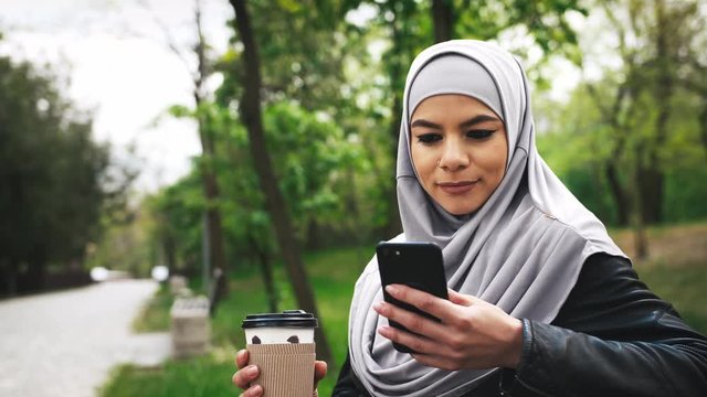 Portrait of young moderm attractive Muslim woman wearing hijab sitting in park and listening to music with smartphone and cup of coffee in her hands