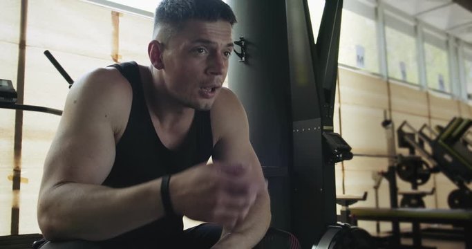 Portrait of professional muscular athletic man bodybuilder in shorts is sitting on bench in closed gym