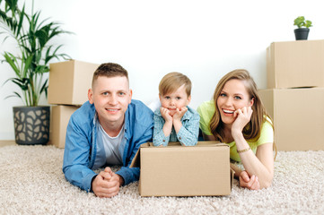 Dreams come true. A young happy family with their little son moved to their new own apartment. They are lying on the floor in an empty apartment among cardboard boxes with things and smiling