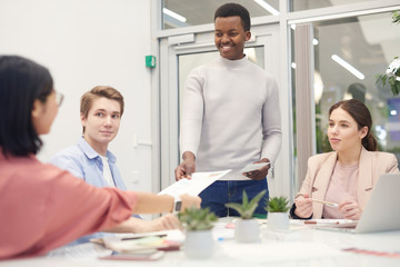 Multi-ethnic group of young business people collaborating on work project during meeting in office, focus on smiling African-American man handing documents