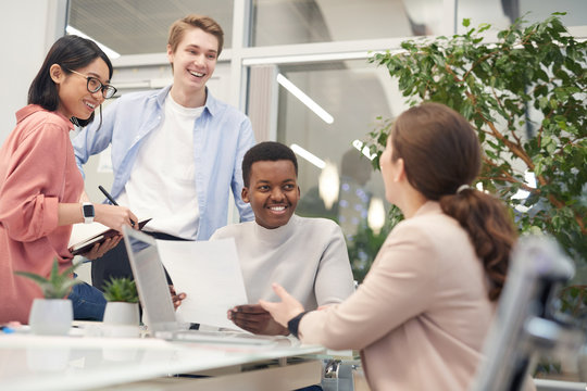 Multi-ethnic group of young business people discussing work project and smiling cheerfully during meeting in office, copy space