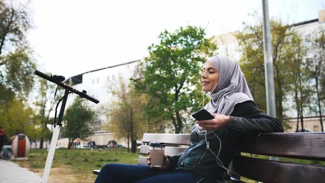 Young moderm attractive Muslim woman sitting in park wearing hijab and listening to music with smartphone and cup of coffee in her hands