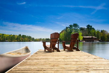 Two Muskoka chairs sitting on a wood dock facing a lake. Across the calm water is a brown cottage...