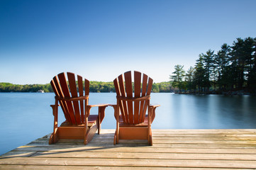 Naklejka premium Two Muskoka chairs sitting on a wood dock facing a lake. Across the calm water are cottages nestled among green trees. There are no clouds in the sky.