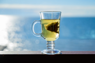 Transparent glass mug with a bag of green leaf tea on a background of the sea.