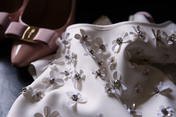 a white dress decorated with flowers made of fabric and pink bride?s shoes stand nearby, details of...