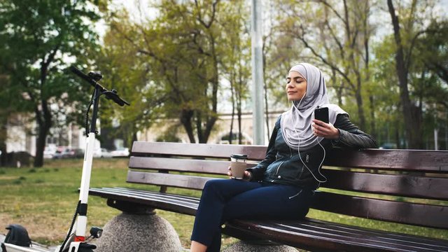 Young moderm attractive Muslim woman sitting in park wearing hijab and listening to music with smartphone and cup of coffee in her hands