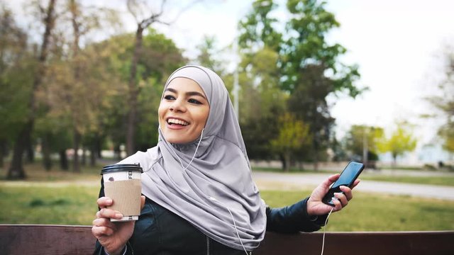 Young moderm attractive Muslim woman sitting in park wearing hijab and listening to music with smartphone and cup of coffee in her hands