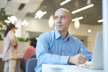 Fototapeta premium Portrait of smiling bald man looking away dreamily while working with laptop in office, copy space