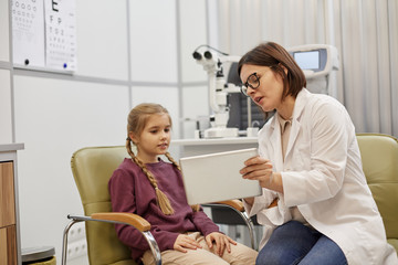 Portrait of young female ophthalmologist using digital tablet while talking to little girl during consultation in modern clinic, copy space