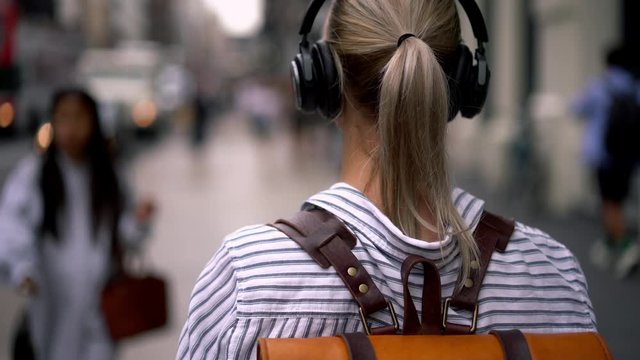 Female tourist with stylish backpack listening music from playlist in modern headphones connected to smartphone device via bluetooth connection.Hipster girl strolling in urban setting
