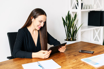 Beautiful woman in black shirt with a digital tablet in hands is sitting at the table in office, watching at tablet screen and smiles. Video meeting, online conversation