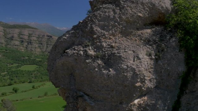 AERIAL DRONE SIDE SHOT DISCOVERING BEHIND ROCKS A BEAUTIFUL GREEN VALLEY IN SPRING WITH SMALL VILLAGE AND PYRENEES IN THE BACKGROUND