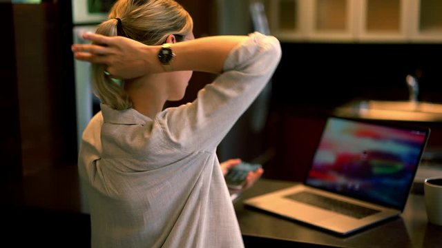 Back view of young woman in eyeglasses holding smartphone in hands and enter pin code on laptop computer for remote work at home.Freelancer checking email on digital netbook
