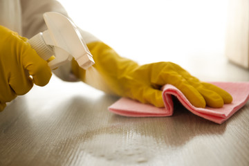 Woman in gloves cleaning wooden table with spray detergent and rag indoors, closeup