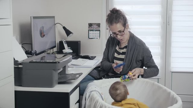 Caucasian working mother stops and plays with infant inside crib in home office