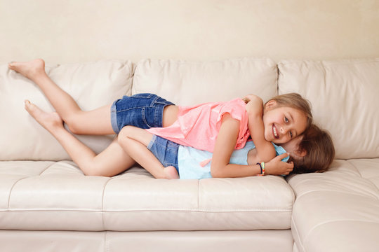 Two cute little Caucasian girls siblings playing at home. Adorable children kids lying hugging on a couch together. Authentic candid lifestyle domestic life moment. Happy friends sisters relationship.