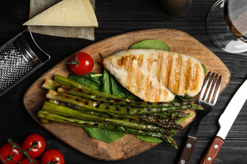 Tasty grilled chicken fillet served with asparagus on black wooden table, flat lay
