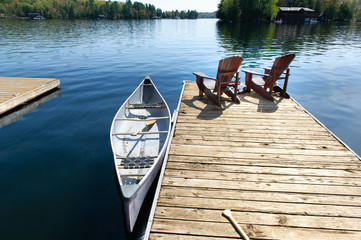 Two Muskoka chairs sitting on a wood dock facing a lake. Across the calm water is a brown cottage...