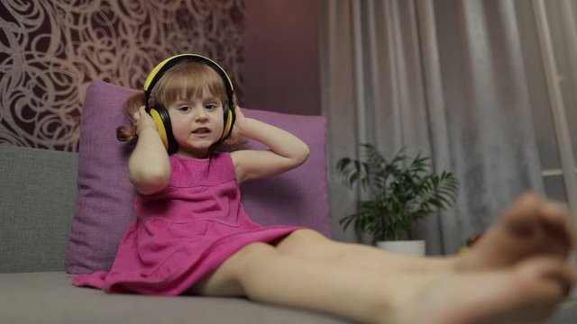 Little child girl in headphones enjoying listen music. Dancing on sofa at home