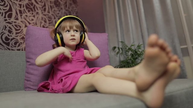 Little child girl in headphones enjoying listen music. Dancing on sofa at home