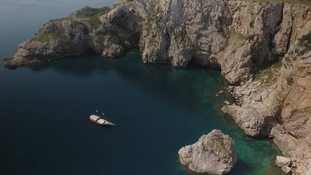 AERIAL DRONE ORBIT OF ANTIC SAIL BOAT ANCHORED IN MEDITERRANEAN COVE, BLUE CALM SEA AND GREEN PINE FOREST