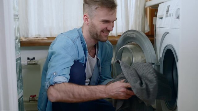 Young bearded man puts clothes in washing machine while sitting on floor in home interior spbas. American guy loads washer and smiling, having nice day in apartment. Hipster is using appliance for
