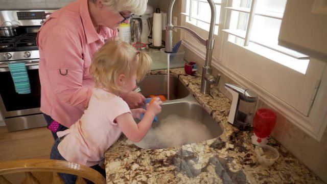 Grandma teaching granddaughter toddler how to wash dishes by washing toys in the kitchen sink