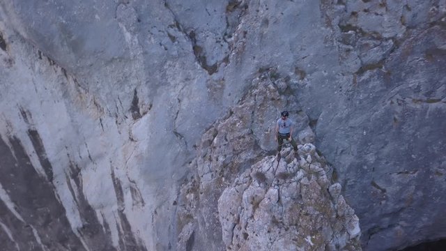 Man in blue shirt and helmet stands on small rocky peak looking out on vast Turda Gorge and deep valley below. Sheer rock walls of terrain. Drone orbiting around rock climber on top of mountain.