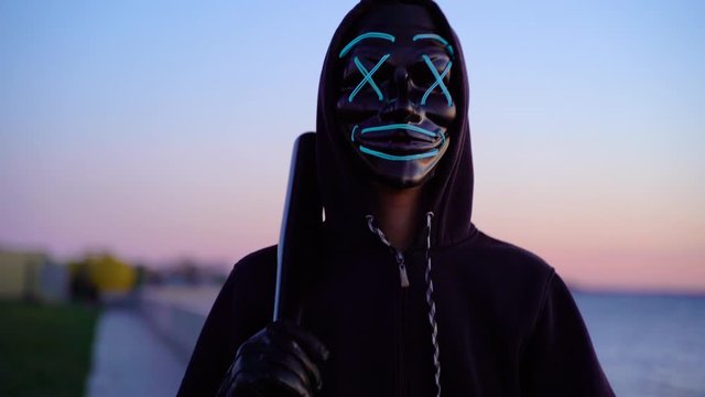 Mysterious Halloween mask. Man in the blue mask glow with a bat in hand at dusk at sunset by the river.