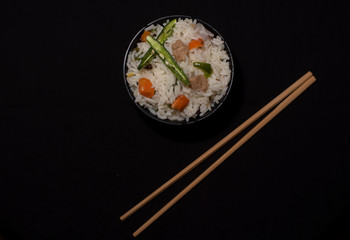 Top down image of a bowl of rice with vegetables and soya chunks along with chopsticks in a black copy space background. Food photography.