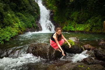 Young Caucasian woman sitting on the rock and playing with water in the river near the waterfall. Travel lifestyle. Dedari waterfall in Sambangan, Bali.