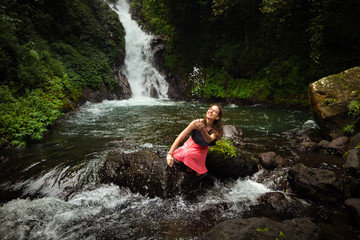 Happy Caucasian woman sitting on the rock and playing with water in the river near the waterfall. Travel lifestyle. Dedari waterfall in Sambangan, Bali.