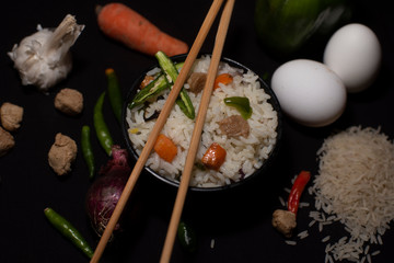 A bowl of a steamed rice and vegetables decorated with veggies, eggs, grains, chopsticks and spoon in a black copy space background. Food photography.