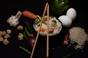 A bowl of a steamed rice and vegetables decorated with veggies, eggs, grains, chopsticks and spoon in a black copy space background. Food photography.