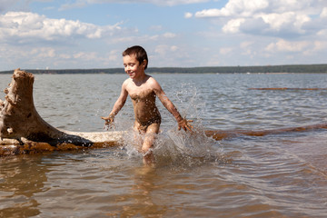 A cheerful little boy enjoys the summer and plays in the water on a warm summer day, splashes from the sea fly in different directions, in the background beach and forest