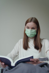 The young beautiful girl found an occupation during quarantine. Girl is reading a book in a medical mask. Orvi, coronovirus, influenza, acute respiratory infections. Vertical photo.