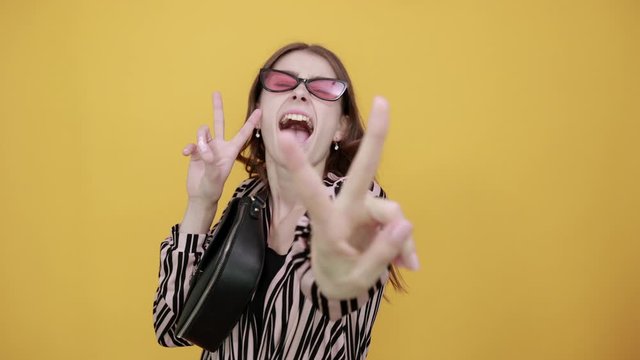 Cute Young Woman Light Brown With Bag In Striped Pink, Black Shirt On A Yellow Background, Expressive Girl Smiles, Has Fun And Shows Peace Sign With Her Fingers