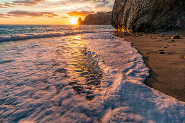 Bright sea sunset. The waves crash into the rock, lit by the warm sunset, sand and pebbles,...