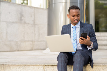 Happy young African businessman working with laptop and phone on the stairs outdoors - obrazy, fototapety, plakaty