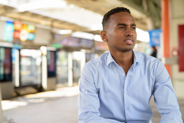 Obraz premium Happy young handsome African businessman thinking while sitting at the sky train station