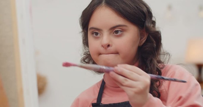 Close up view of beautiful teenager girl with genetic disorder sitting in front of molbert. Nice looking young ladywith brown wavy hair holding paintbrush and creating picture. Indoors.