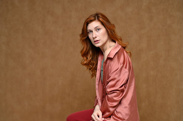 Fototapeta premium Photo of a young caucasian red-haired woman on a beige background. A pretty model with long hair is dressed in red trousers and a pink blouse.