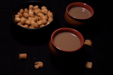 Hot infused tea in earthen cups along with a bowl of snacks kept in black copy space background. Indian beverages and food photography.