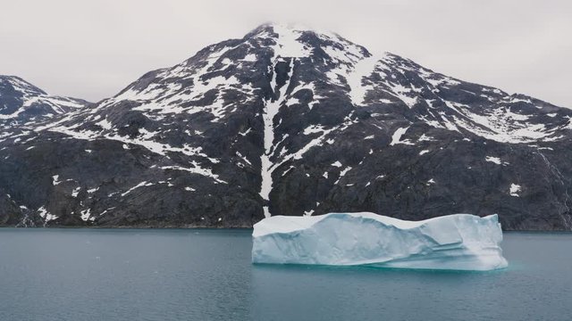 Iceberg floating surrounded by snow capped mountains in Prince Christian Sound in Greenland filmed from the side of a cruise ship in slow motion