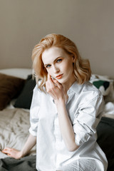 A young beautiful woman in a white shirt wakes up in the morning, sitting on the bed. A pretty woman keeps croissants and a Cup of hot drink in the bedroom. The concept of food and way of life.